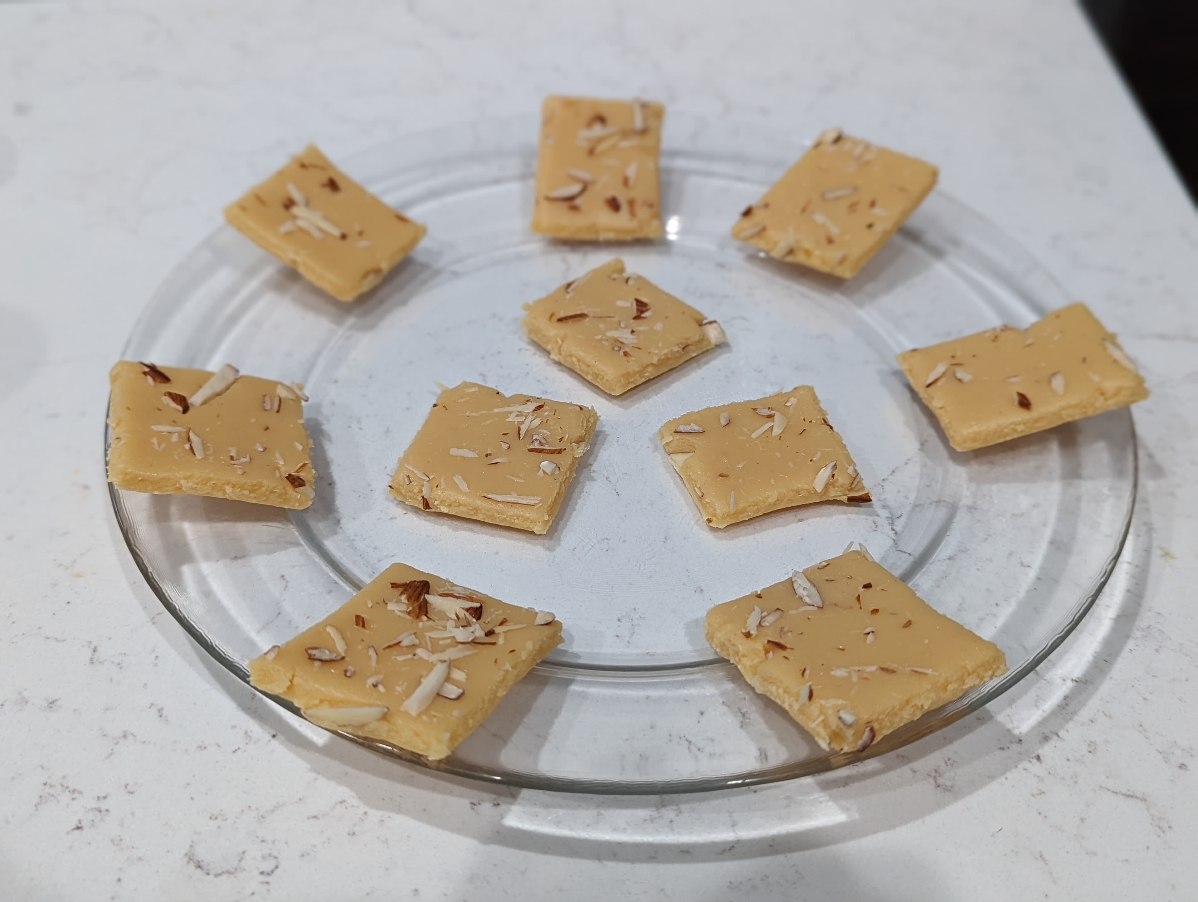 Square pieces of milk burfi topped with almond slivers on a glass plate