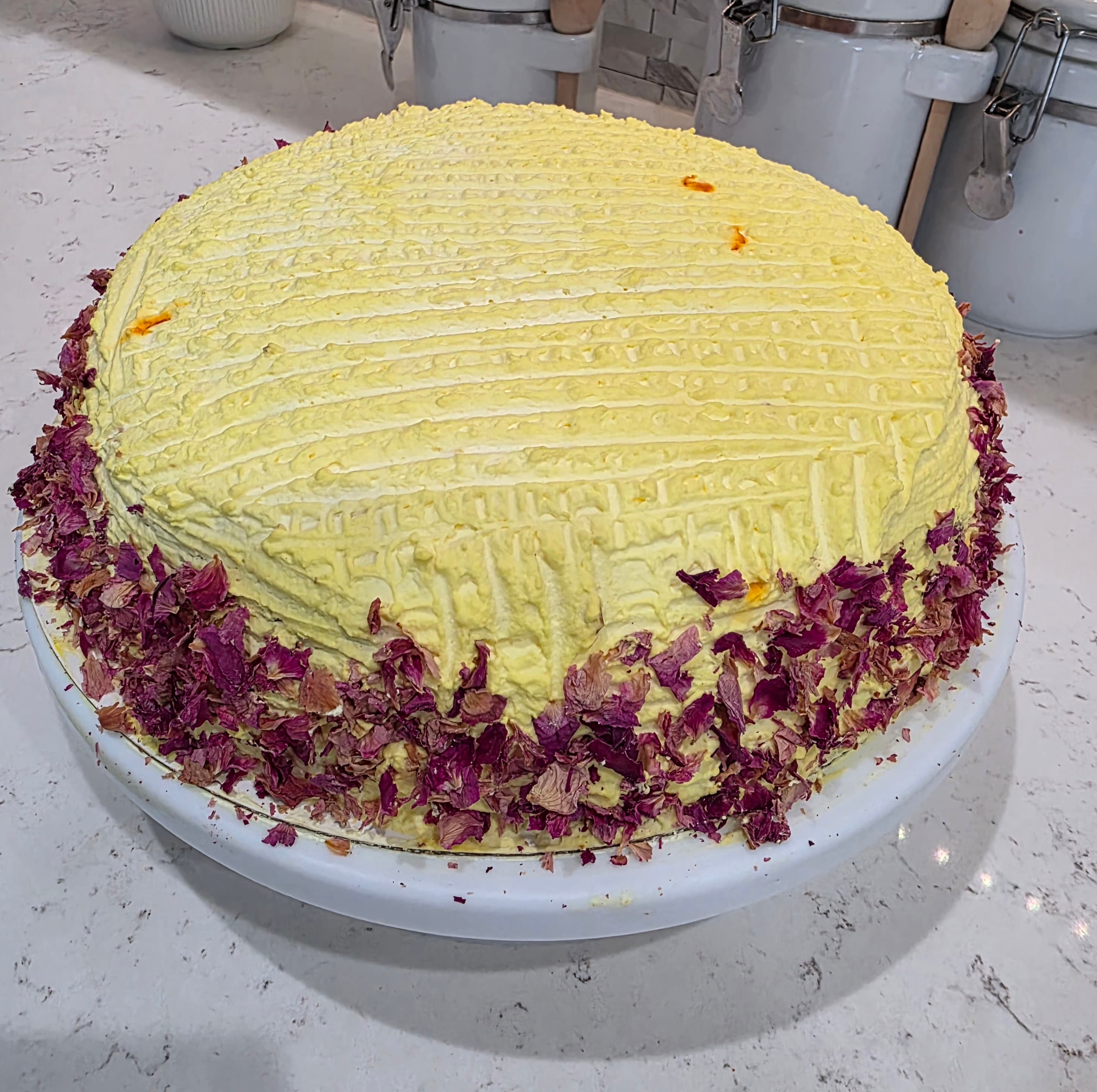 Homemade rasmalai cake with saffron frosting and dried rose petals on a white cake stand
